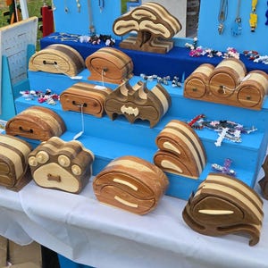May include: A display of handcrafted wooden boxes in various shapes, including a paw print, a bat, and a tree. The boxes are made of light and dark wood, with drawers and decorative elements. The display also includes necklaces and other small accessories.