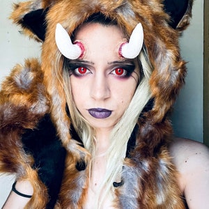 May include: A person wearing a brown and grey faux fur hooded jacket with white horns and red contact lenses.