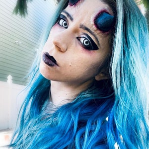 May include: A woman with blue hair and white contact lenses is wearing black eyeliner and dark lipstick. She has fake horns on her forehead.