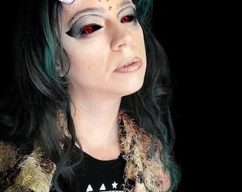 May include: A woman with green hair and white horns is wearing red contact lenses and a black t-shirt with the text "We live by the", a gold crescent moon and stars on her forehead, and a brown and gold fuzzy shawl.