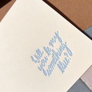 May include: A cream-coloured card with the light blue text "Will be my something blue?" in a handwritten style. The card is angled, revealing a glimpse of a brown envelope and a grey surface.