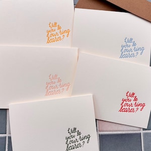 Peut inclure: Cartes blanc cassé avec l'inscription "Will you be your ring bearer ?" en orange, bleu, rose, rouge et noir. Une enveloppe marron est visible en arrière-plan. Les cartes sont disposées sur une surface carrelée.