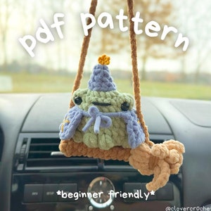 Könnte beinhalten: Ein gehäkelter Frosch-Autoanhänger mit Partyhut und Umhang. Der Frosch ist grün, mit blauem Hut und Umhang und einem braunen Sockel. Der Text "pdf pattern" und "beginner friendly" sind sichtbar.
