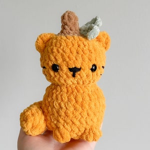 Può includere: Un giocattolo di peluche a forma di zucca arancione lavorato a uncinetto con una foglia verde in cima. Il giocattolo ha occhi neri e un sorriso cucito.