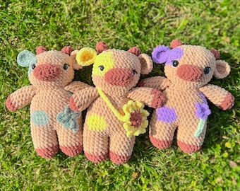 Puede incluir: Tres animales de peluche de crochet con forma de vaca. Las vacas son marrones con detalles coloridos. Una vaca tiene una flor amarilla, otra una flor morada y la última una flor azul.