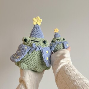May include: Two crocheted frog plushies wearing purple and yellow party hats. The larger frog is wearing a purple cape with yellow stars.