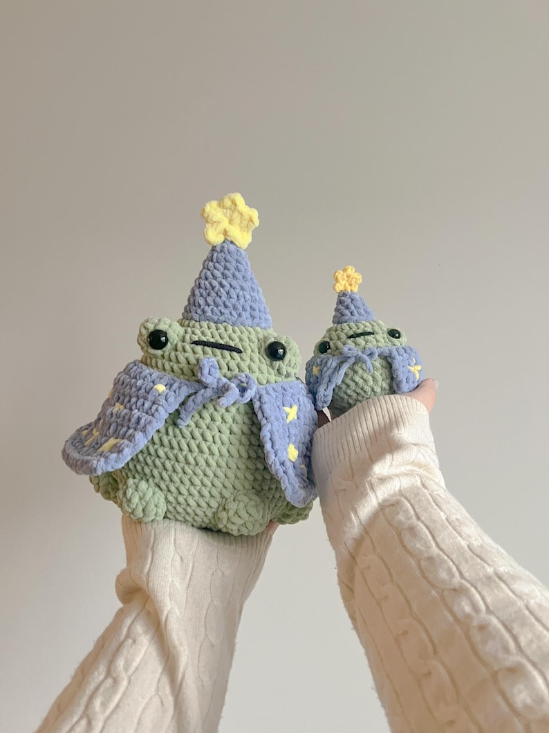 May include: Two crocheted frog plushies wearing purple and yellow party hats. The larger frog is wearing a purple cape with yellow stars.