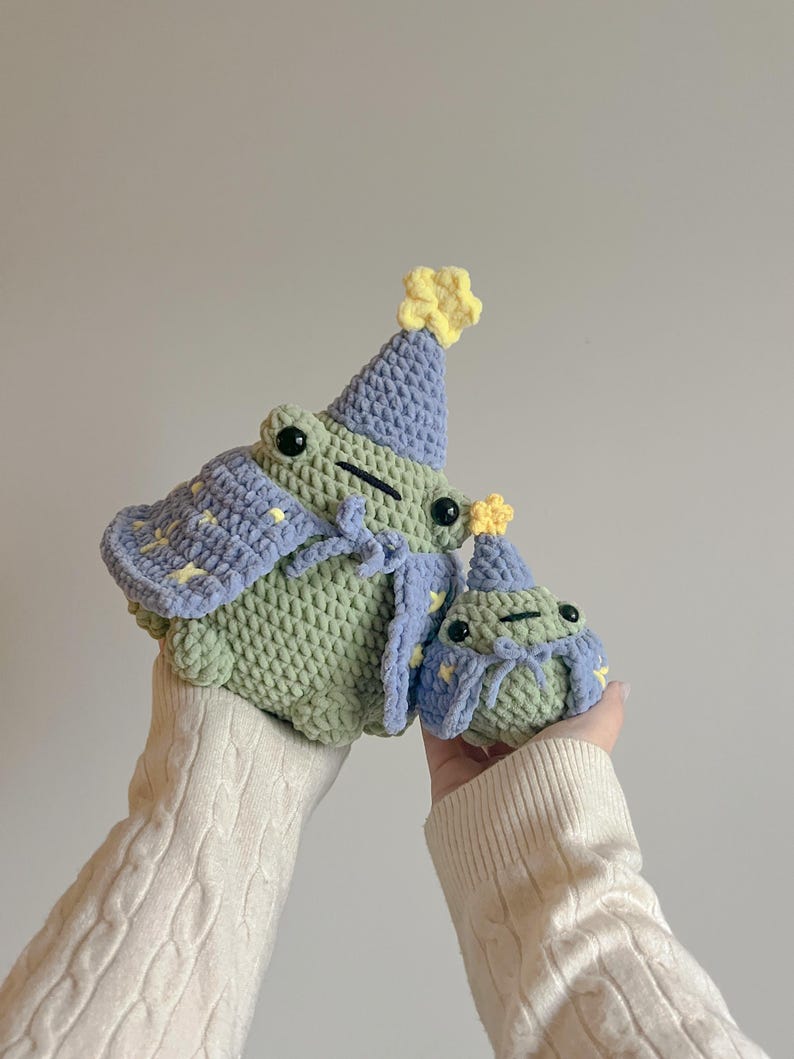 May include: Two crocheted frog plushies wearing party hats. The larger frog is green with a purple and yellow star patterned cape. The smaller frog is green with a purple and yellow star patterned hat.