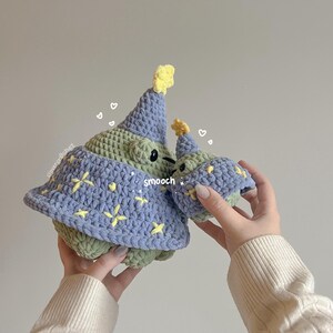 May include: Two crocheted plush toys shaped like a creature wearing a party hat. The larger toy is green and blue with yellow stars. The smaller toy is green with a yellow star on its hat. The text "smooch" is written on the larger toy.