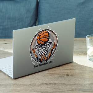 以下が含まれることがあります： バスケットボールの漫画のキャラクターがバスケットボールをゴールにダンクシュートしているステッカーが貼られたシルバーのラップトップ。ステッカーには「Since December 1891」という文字が書かれている。