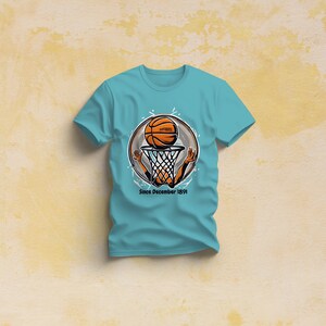 以下が含まれることがあります： ターコイズブルーのTシャツに、バスケットボールがフープを通過するグラフィックがプリントされています。グラフィックは白地に黒のアウトラインで描かれた円の中にあります。グラフィックの下には「Since December 1991」というテキストが書かれています。