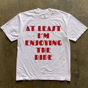 May include: White t-shirt with red text that reads "At Least I'm Enjoying The Ride".