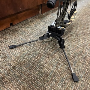 May include: A compound bow with an olive green riser and camouflage limbs, supported by a black folding tripod stand. The bow features a Trophy Ridge stabilizer attached to the front, with black cables and cams visible in the background structure.