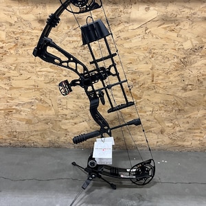 May include: A black compound bow with a sight, stabilizer, and arrow rest. The bow is set on a stand. The brand name "BOWTECH" is visible on a tag attached to the bow.