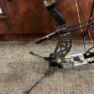 May include: An olive green compound bow with black accents and a Trophy Ridge stabilizer. The bow features a sight with yellow and black components, and an orange arrow. The bow is set on a stand on a patterned carpet.