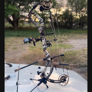 May include: A compound bow with a camouflage pattern, featuring a sight, stabilizer, and arrow rest. The bow is set up on a white surface, with a tripod stand. The background shows a natural outdoor setting.