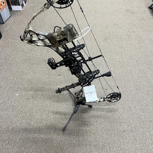 May include: A compound bow featuring a camouflage pattern finish, black cams, and a black cable system. The bow is equipped with a black quiver, a sight, and a stabilizer. A white tag with the text BOWTECH is attached to the riser of the bow.
