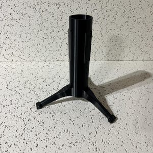 May include: A black, cylindrical stand featuring a hollow vertical tube supported by three radiating, angled legs. The structure is composed of a smooth, matte material and is designed to provide a stable base for upright objects or components.