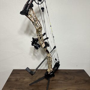 May include: A camouflage-patterned compound bow with black accents and a black bow stand. The bow is strung and has a sight and a rest.