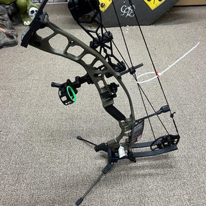 May include: An olive green compound bow featuring a black riser, a multi-pin sight with a bright green circular housing, black cams, and a black string system. The bow is mounted on a black tripod stand and includes a white zip tie attached to the cables.