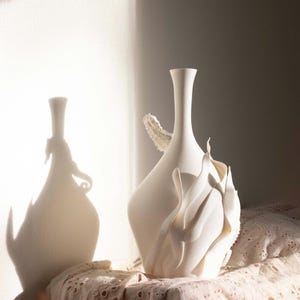 May include: Two white ceramic vases with unique designs sit on a lace-covered surface. One vase has a tall, slender neck, while the other features an organic, sculptural form. The sunlight casts dramatic shadows on the wall behind them.