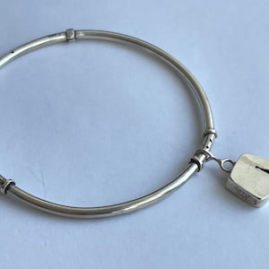 May include: A silver bangle bracelet with a square charm. The bracelet has a circular design with a hinge and a small square charm with a keyhole design. The bracelet is made of a shiny metal.