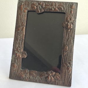 May include: Bronze-colored picture frame with floral and leaf detailing. The frame has a rectangular shape with a dark interior, and the design includes raised iris flowers and foliage. The frame is standing upright on a white surface.