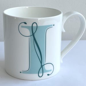 May include: A white ceramic mug featuring a large, light blue capital letter I decorated with dark teal, swirling calligraphic flourishes. The mug has a smooth, glossy finish and a curved handle on the right side, set against a plain background.