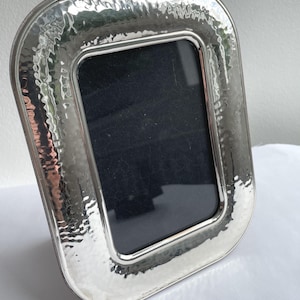 May include: A rectangular silver-toned picture frame with rounded corners and a distinct hammered texture on the wide border. The frame features a smooth inner edge surrounding a dark, solid-coloured rectangular insert, standing upright on a surface.