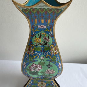May include: A decorative, ornate vase with a flared top and a wooden base. The vase features a blue background with floral and bird designs in shades of pink, green, and yellow, accented with gold trim.
