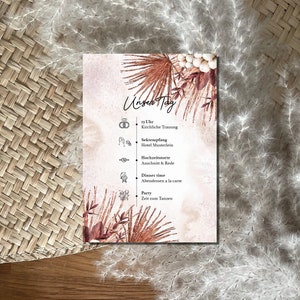 Könnte beinhalten: Eine hellrosa Hochzeitseinladungskarte mit floralem Design. Die Karte listet den Zeitplan für den Hochzeitstag auf, einschließlich der Trauung, des Empfangs, des Abendessens und der Party. Der Text auf der Karte lautet "Unser Tag".