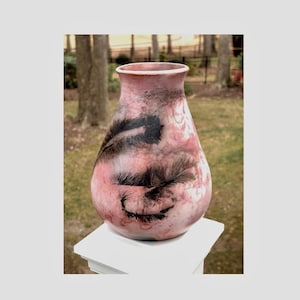 May include: A decorative vase with a pink and white marbled finish. The vase features black feather-like designs. The vase is displayed on a white surface, with a blurred outdoor background of trees and grass.