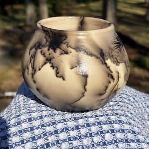 May include: A handmade ceramic bowl with a rounded shape and a wide opening. The bowl has a cream-colored base with dark, lightning-like patterns. The bowl is sitting on a blue and white patterned cloth.