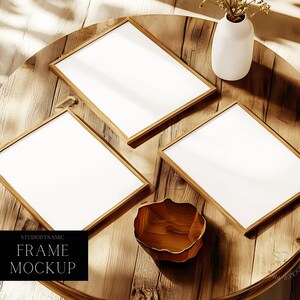 Puede incluir: Tres marcos de fotos dorados con paspartús blancos sobre una mesa de madera con un jarrón blanco y flores secas. Los marcos están vacíos y listos para tus fotos. Studiodynamic Frame Mockup.