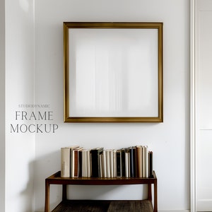 May include: A gold picture frame with a white background hangs on a white wall. A wooden shelf with a row of books sits below the frame.