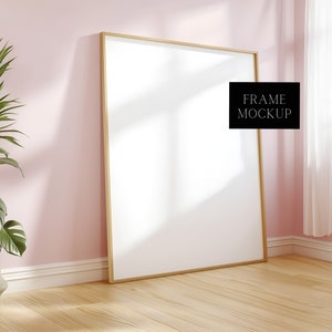 May include: A large, empty, wooden frame with a white background, leaning against a pink wall. The frame is in a room with a wooden floor and a white curtain in the background. The text "FRAME MOCKUP" is in a black box in the upper right corner of the frame.