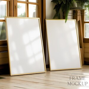 May include: Two gold-framed picture frames are leaning against a wooden wall. The frames are empty and have a white background. The frames are in a room with a wooden floor and a window in the background.