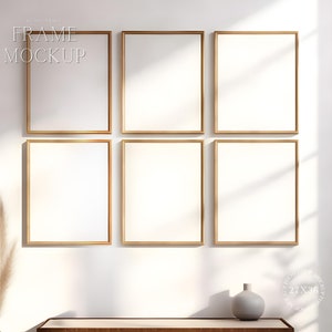 May include: Six empty picture frames with gold trim are arranged in a grid on a white wall. The frames are all the same size and are evenly spaced. The wall is painted a light cream color and there is a wooden table in the foreground.