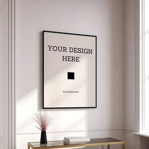 Pu&ograve; includere: Un poster incorniciato nero con il testo "YOUR DESIGN HERE" e le parole "StudioDynamic" in basso. Il poster &egrave; appeso a una parete bianca in una stanza con una finestra e un tavolo.