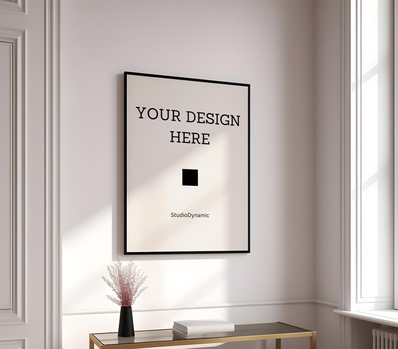 Pu&ograve; includere: Un poster incorniciato nero con il testo "YOUR DESIGN HERE" e le parole "StudioDynamic" in basso. Il poster &egrave; appeso a una parete bianca in una stanza con una finestra e un tavolo.