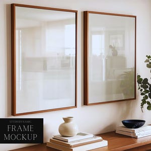 May include: Two rectangular wooden framed art mockups hanging on a white wall. The frames have a light brown wood finish. A black sign at the bottom left reads "STUDIODYNAMIC FRAME MOCKUP". A vase, books, and a bowl are on a wooden table.