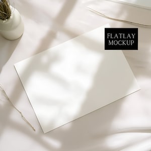 May include: A flatlay mockup featuring a blank white paper, a small white vase with dried grass, and a shadow overlay. The text "FLATLAY MOCKUP" is in a black rectangle. The scene is set on a white, textured surface.