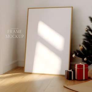 Könnte beinhalten: Ein großer, leerer Bilderrahmen in Gold steht in einer Ecke eines Raumes mit Holzboden. Im Vordergrund steht ein kleiner Weihnachtsbaum mit goldenen Kugeln und zwei verpackte Geschenke.