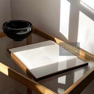 Puede incluir: Una maqueta de plano plano con una imagen enmarcada en blanco sobre una bandeja de madera con un tazón negro. La bandeja está sobre una superficie de vidrio con la luz del sol entrando por una ventana.