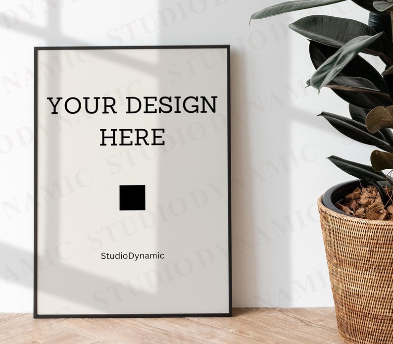 May include: A black framed poster with the text "YOUR DESIGN HERE" and the words "StudioDynamic" at the bottom. The poster is hanging on a white wall with a potted plant in the corner.