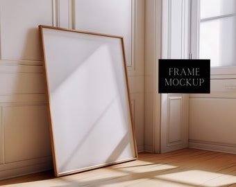 May include: A large, empty, white picture frame with a natural wood frame sits on a wooden floor in a room with white walls and a window. The text "FRAME MOCKUP" is visible in the image.