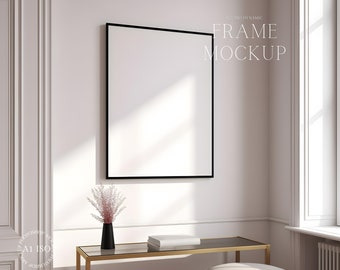 May include: A black picture frame mockup hanging on a white wall with a gold table and a vase with pink flowers in front of it.