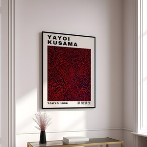 Pu&ograve; includere: Un poster in bianco e nero con il testo "Yayoi Kusama" in grassetto, lettere nere. Il poster presenta un motivo astratto rosso e nero di cerchi e il testo "Tokyo 1998" e "草間彌生" in lettere nere.