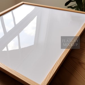 以下が含まれることがあります： 白い背景の木製フレーム。フレームは木製の表面の上に置かれています。フレームの右下隅に「FLATLAY MOCKUP」というテキストが表示されています。