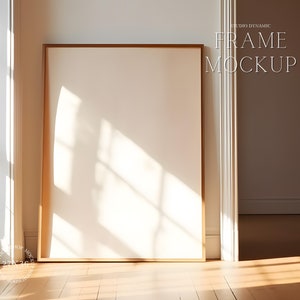 Könnte beinhalten: Ein heller Holzrahmen mit einem weißen Passepartout und einer leeren weißen Leinwand im Inneren. Der Rahmen steht auf einem hellen Holzboden mit einer weißen Wand dahinter. Der Rahmen befindet sich in einem Raum mit einem Fenster links und einer weißen Tür rechts. Der Text "FRAME MOCKUP" ist in Weiß an der Wand geschrieben.
